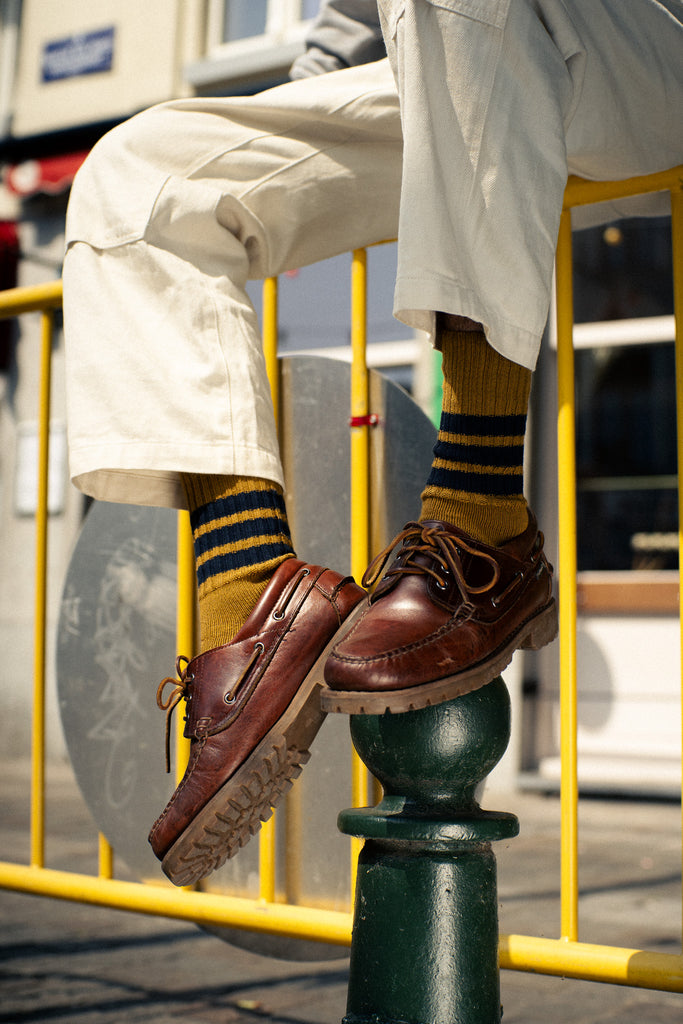 Person wearing brown shoes and yellow socks with blue stripes, sitting on a green metal post.