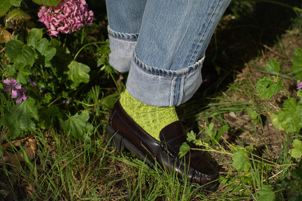 Person wearing green socks and black shoes with rolled-up jeans standing in a garden.