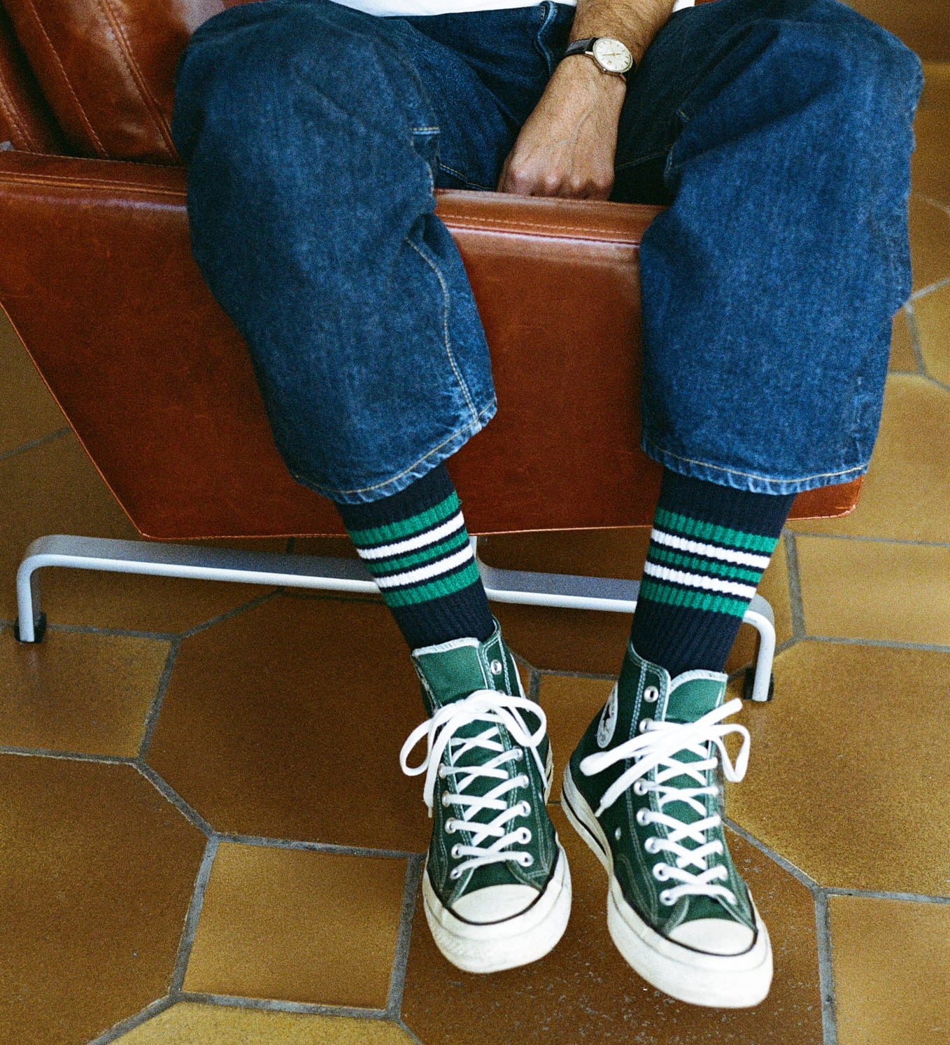 Person wearing green sneakers and blue jeans sitting on a brown leather chair with a tiled floor.