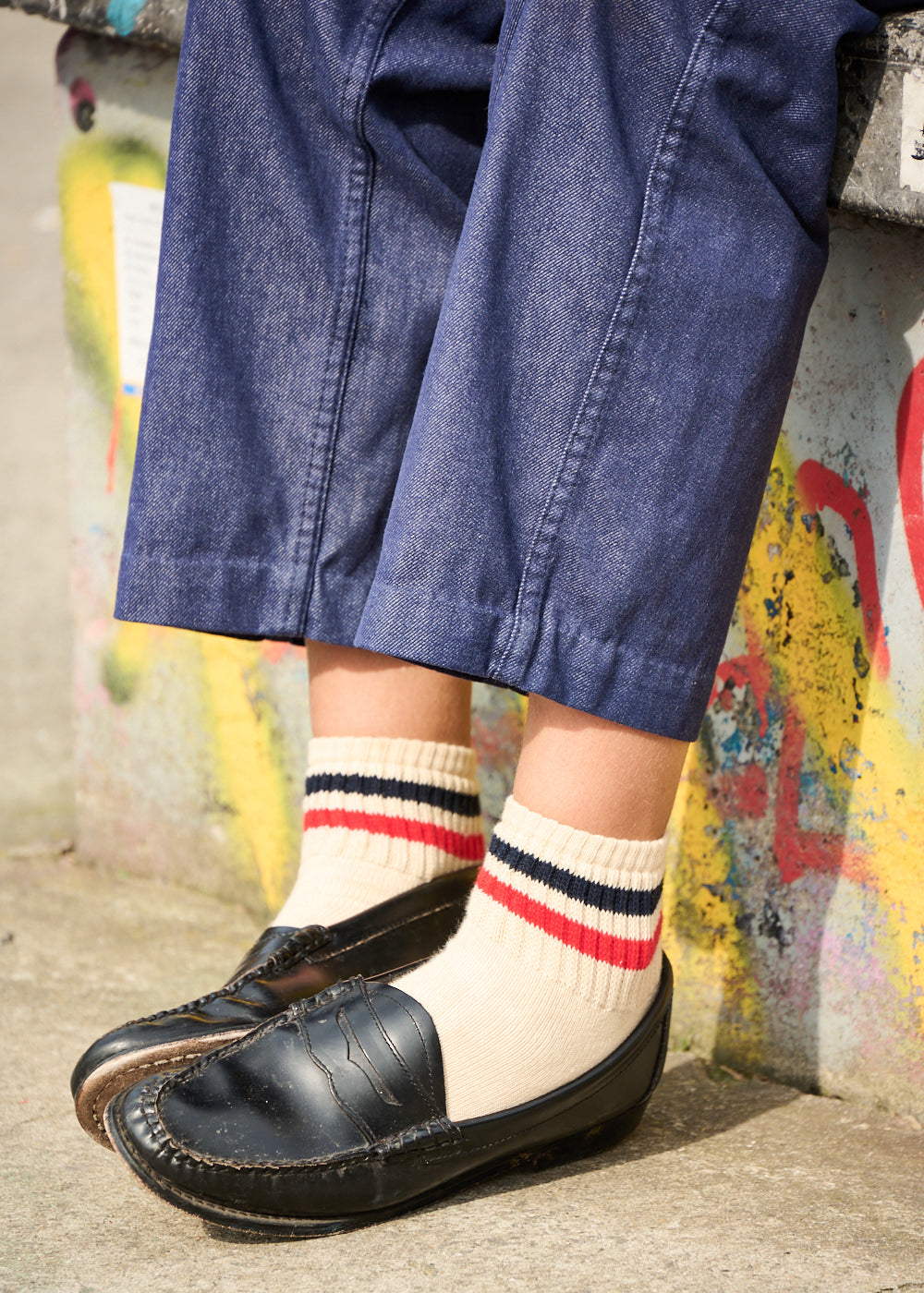 Person wearing black loafers and colorful striped socks with blue jeans against a graffiti-covered wall.