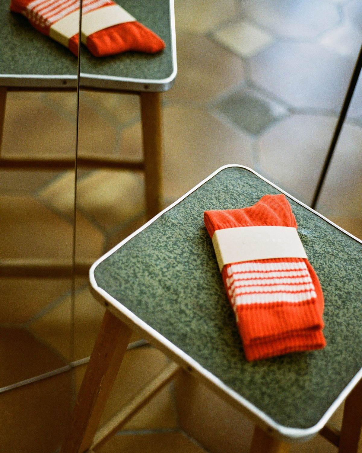 A pair of folded orange knit socks with white stripes sits on a green textured stool with a mirror reflecting the scene.